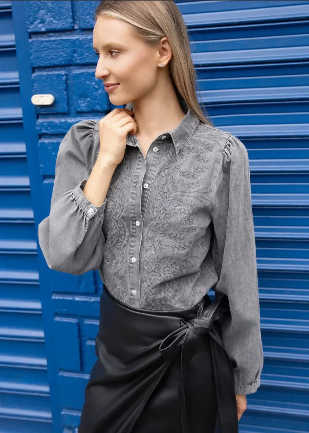 Femme portant une chemise grise à broderie et jupe portefeuille noire, fond bleu urbain