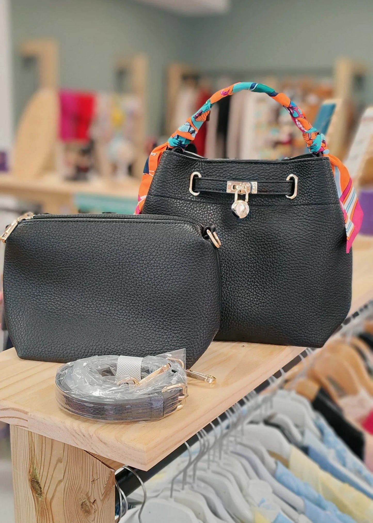 Sac à main noir en simili cuir avec anse ruban colorée, pochette assortie et bandoulière, posé sur une étagère en boutique.