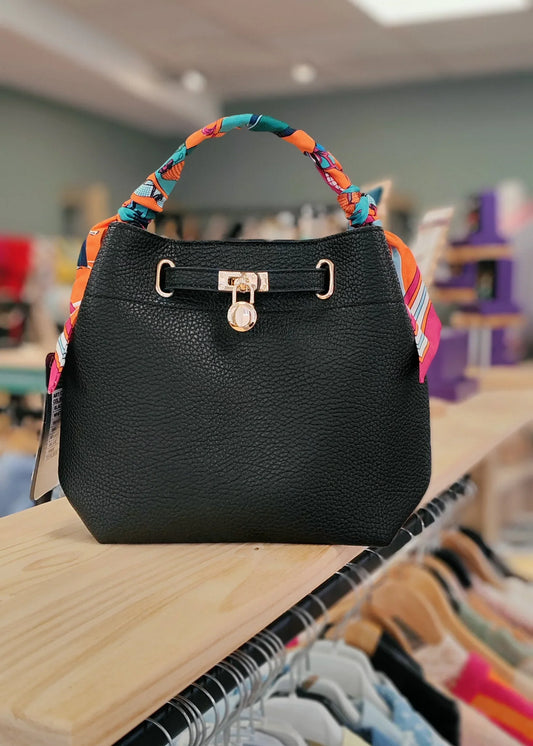 Sac à main noir femme avec poignée ruban coloré et fermoir doré, posé sur étagère.