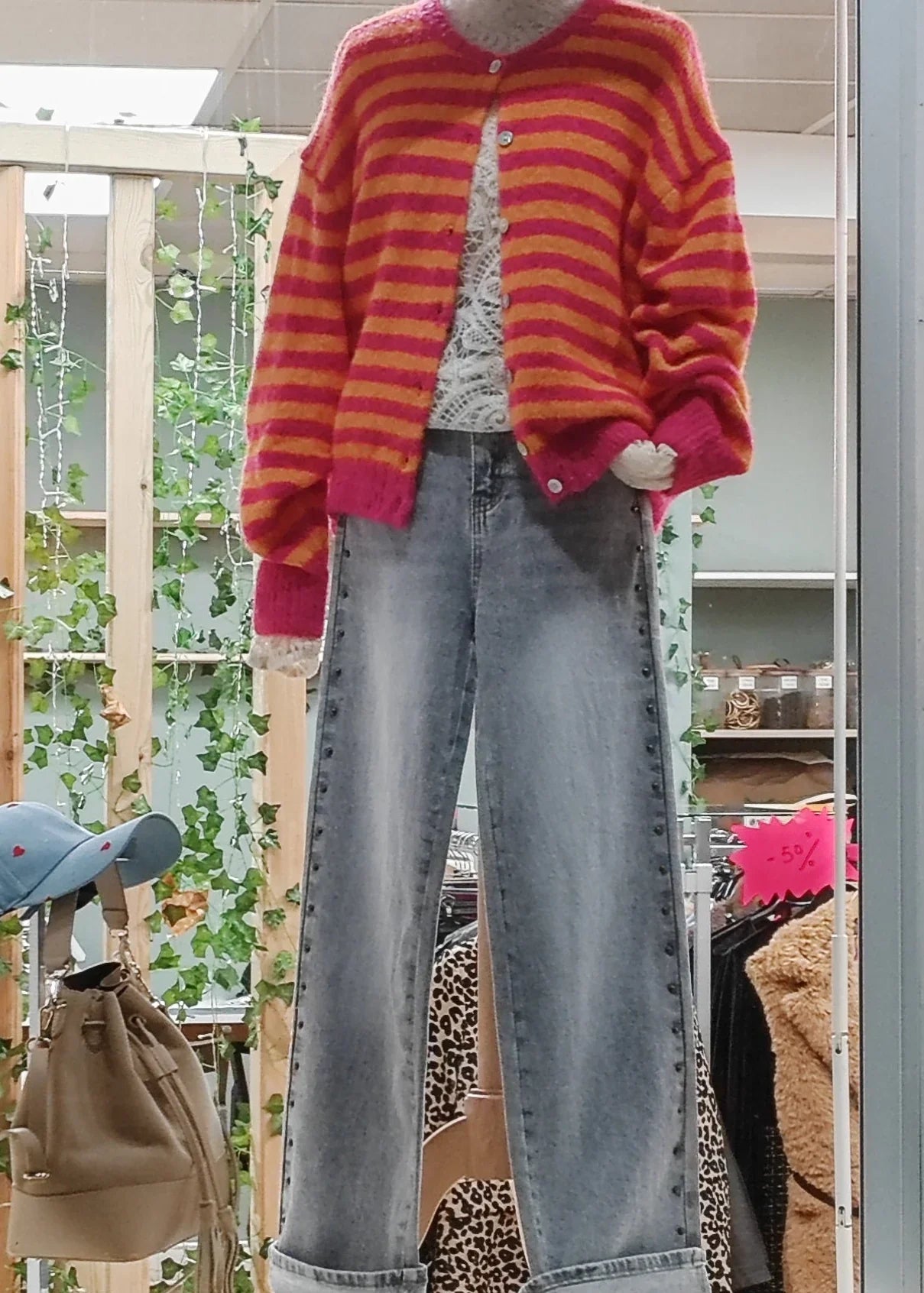 Mannequin avec gilet rayé rose et orange, top dentelle blanc et jean large délavé, devant décor végétal.